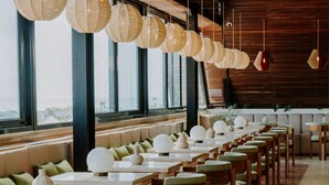 Un restaurante moderno con mesas largas y sillas acolchadas verdes, ventanas amplias y lámparas colgantes esféricas.