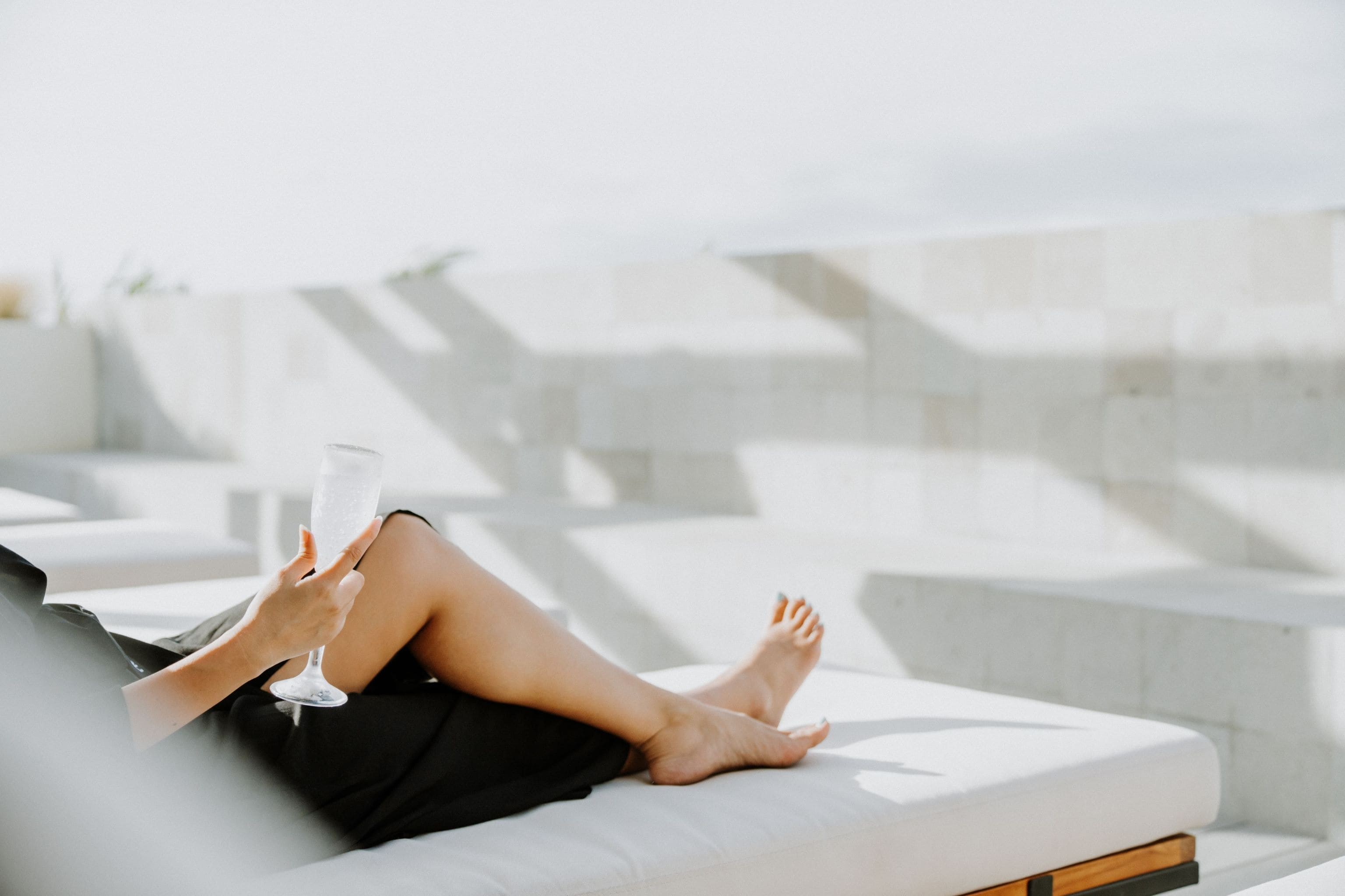 A person reclining on a white outdoor sofa with a glass of drink, set against a modern architectural backdrop.