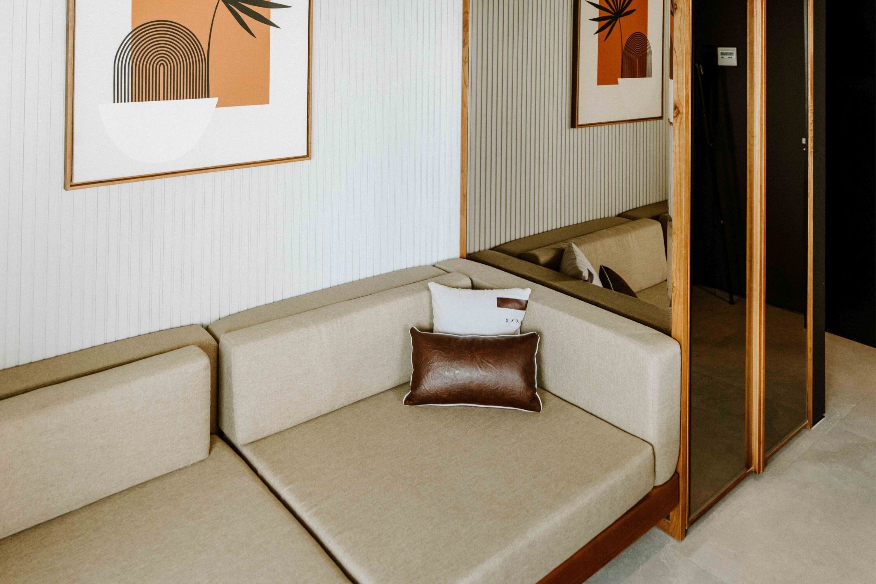 A modern hotel room with a beige sofa, a brown leather pillow, and framed artwork on the wall.