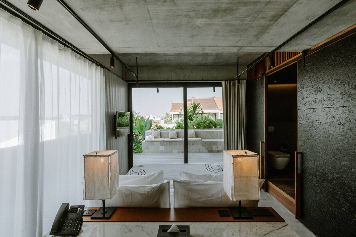 Una habitación de hotel moderna con un ventanal grande, un sofá, una mesa con una lámpara y un armario de madera.