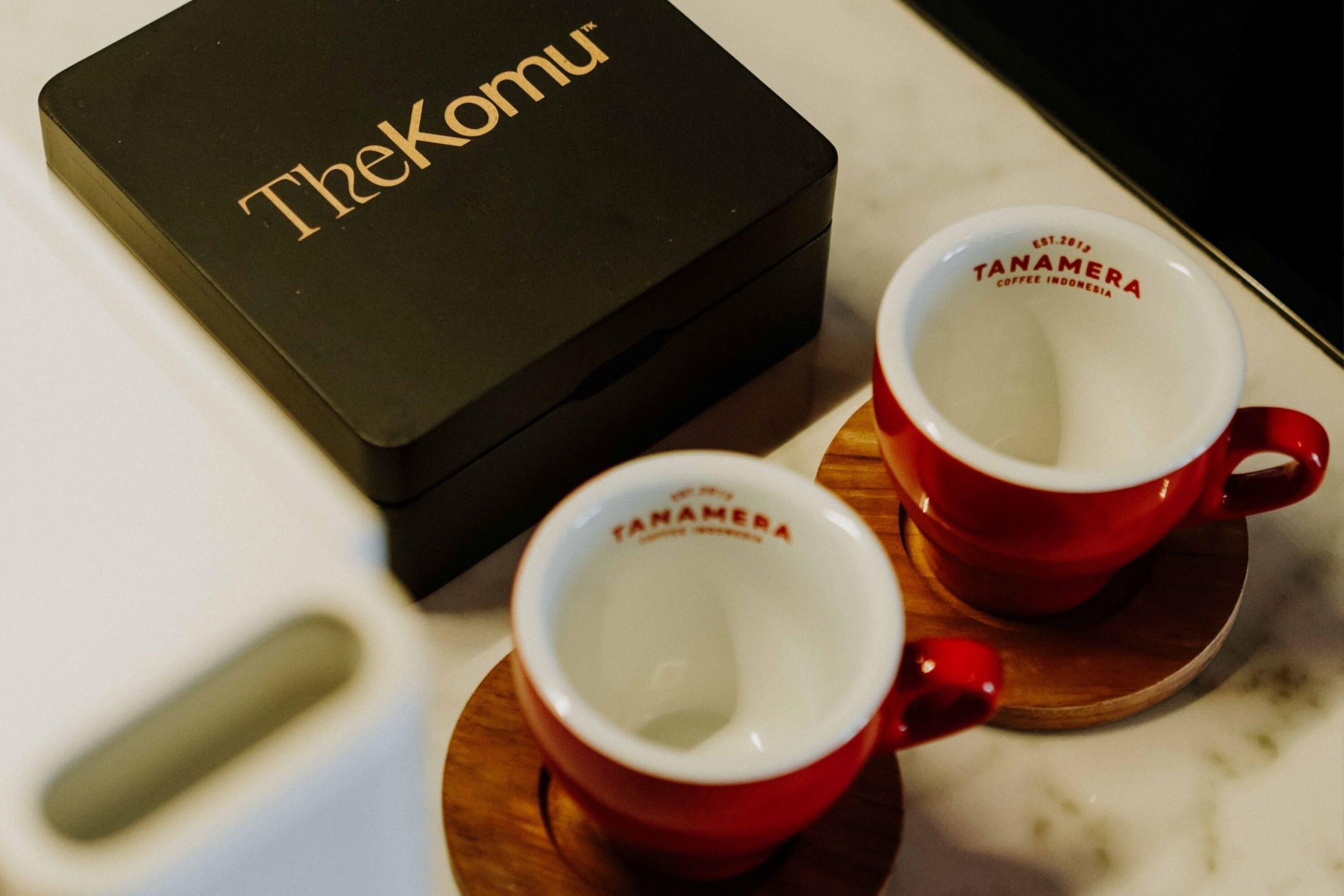A black box with "TheKomu" text, two red cups with "Tanamera Coffee Indonesia" on them, and a wooden coaster.