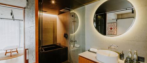 A modern bathroom with a glass-enclosed shower, a freestanding bathtub, a round mirror, and a wooden vanity.