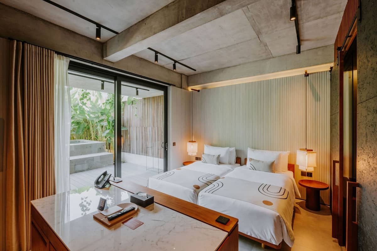 Una habitación de hotel con un gran ventanal corredero que da a un balcón, dos camas con ropa blanca, una mesita de noche de mármol y otra mesita de noche pequeña con una lámpara.
