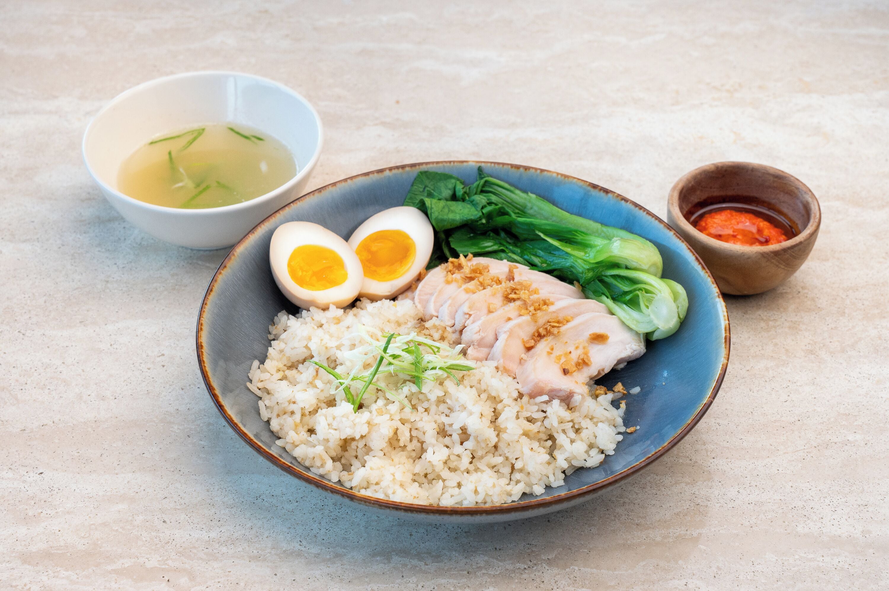 A bowl of rice with sliced chicken, boiled eggs, and bok choy, accompanied by a small bowl of soup and a container of sauce.