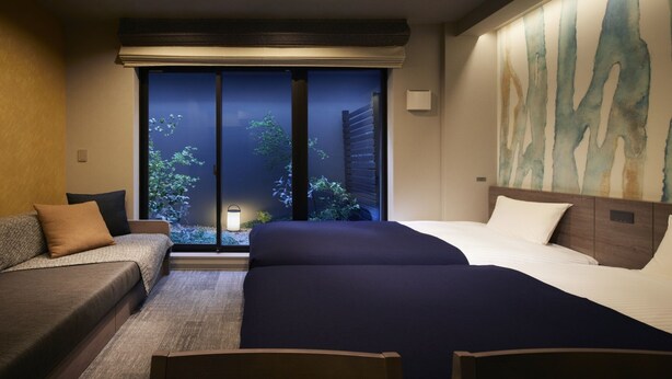 Habitación de hotel con cama, sofá y vista a la vegetación a través de una ventana.