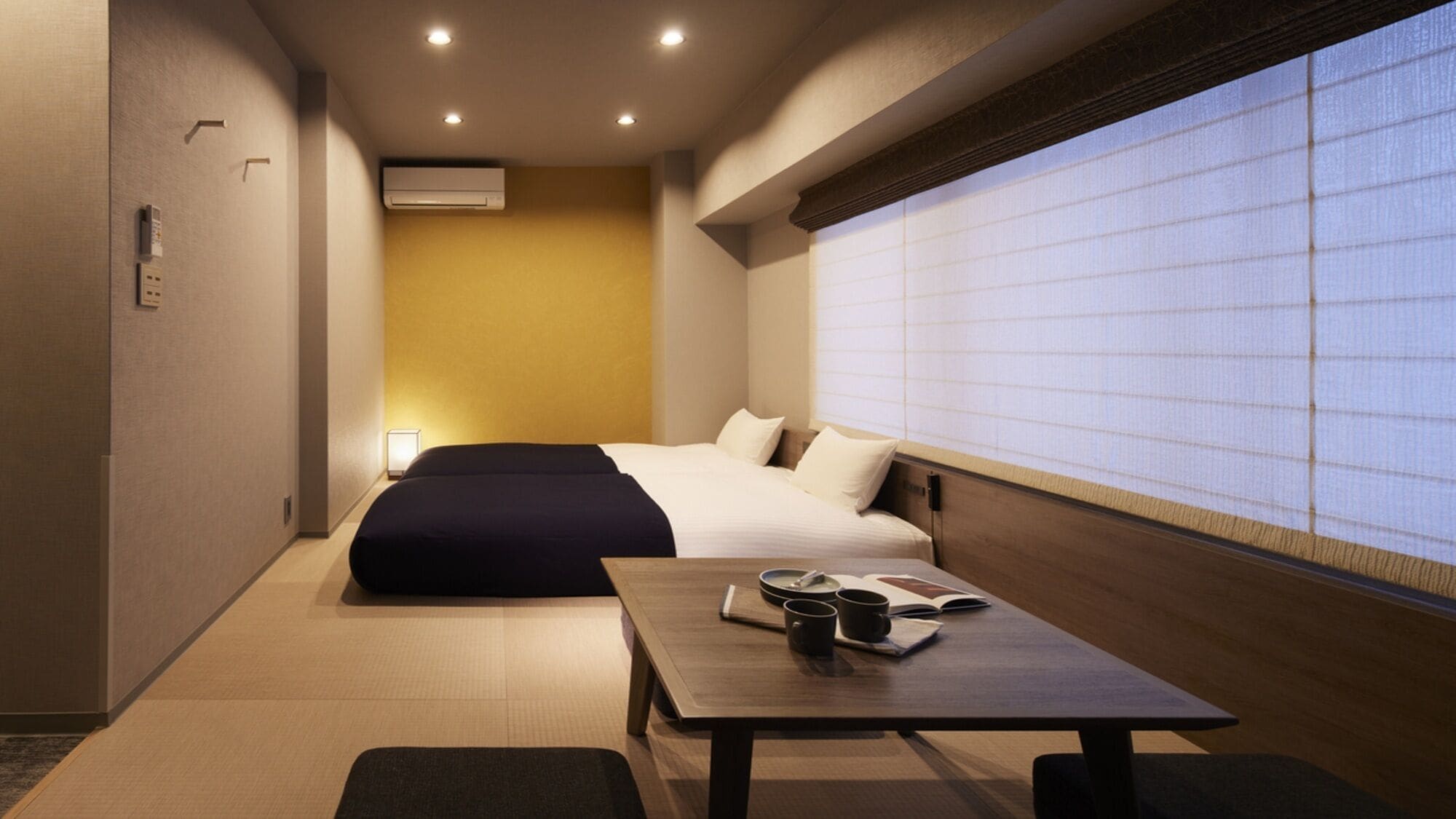 A hotel room with a bed, a table with a book and a cup, and a window with a view.