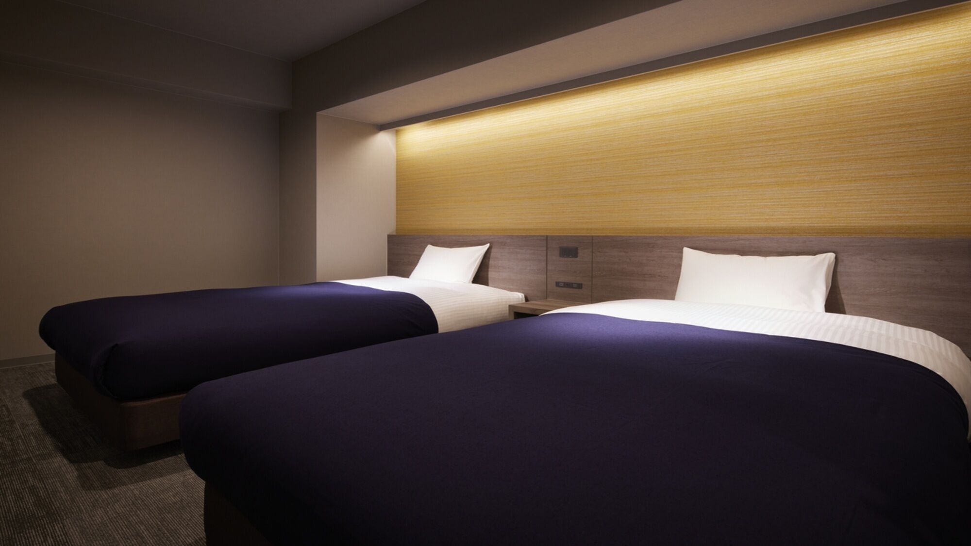 A hotel room with two beds, each with a wooden headboard and a light-colored wall behind.