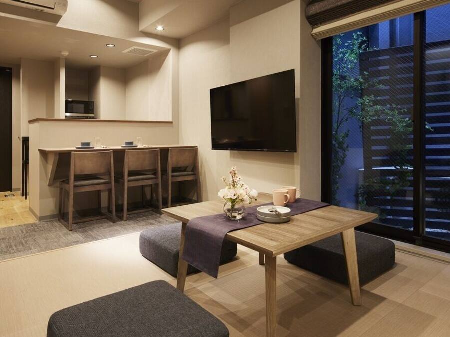 A modern hotel room with a dining area, a TV mounted on the wall, and a large window with a view of the outdoors.