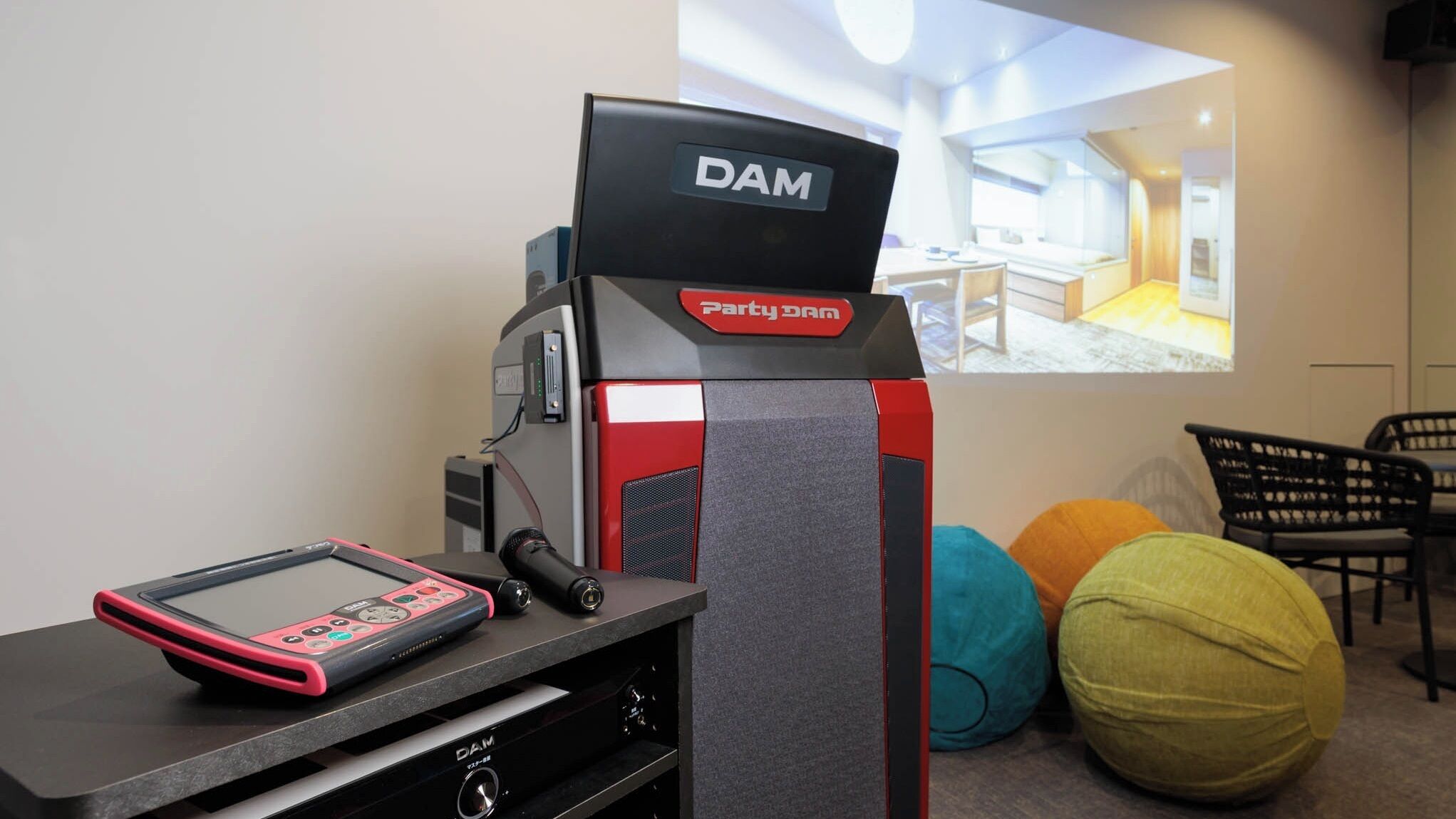 A DAM branded device with a remote control, a projection screen displaying a room, and colorful bean bags in the background.