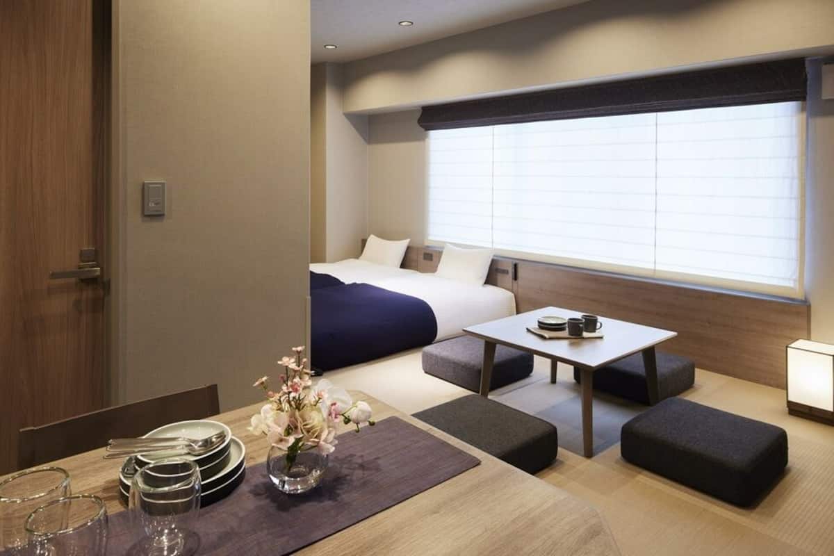 A compact hotel room with a bed, a small table, a bedside table, a window with blinds, a bedside lamp, and a flower vase.