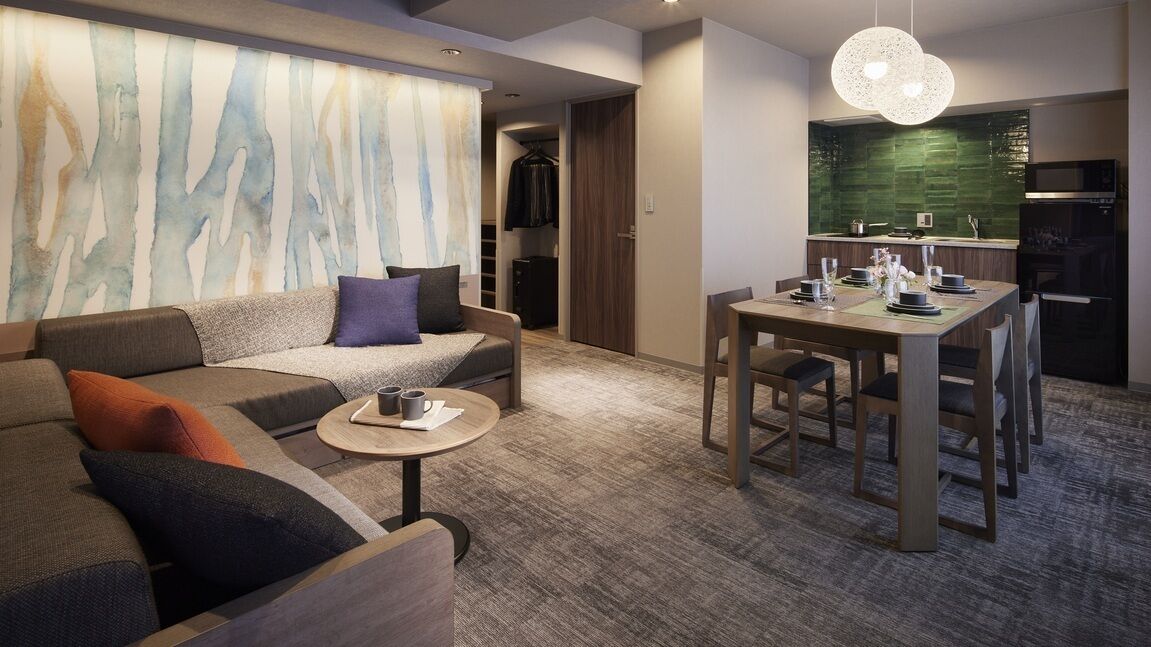 A modern hotel room with a sofa, dining table, and a kitchen area.