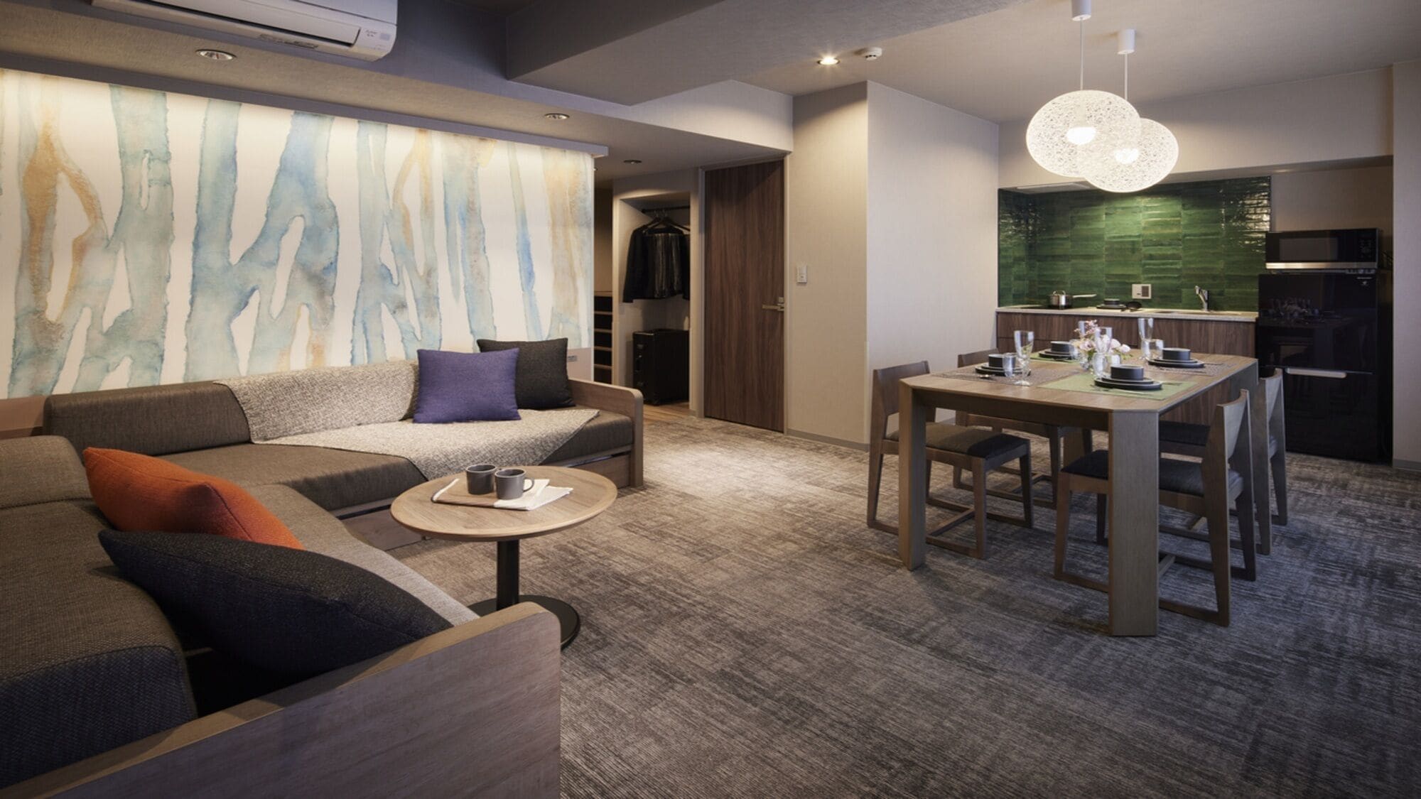 A modern hotel room with a sofa, a small round table, a dining area with a table and chairs, and a kitchen area with a microwave and a green backsplash.