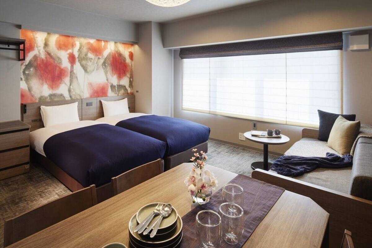 A hotel room with two beds, a sofa, a dining table with a vase of flowers, and a large window with blinds.
