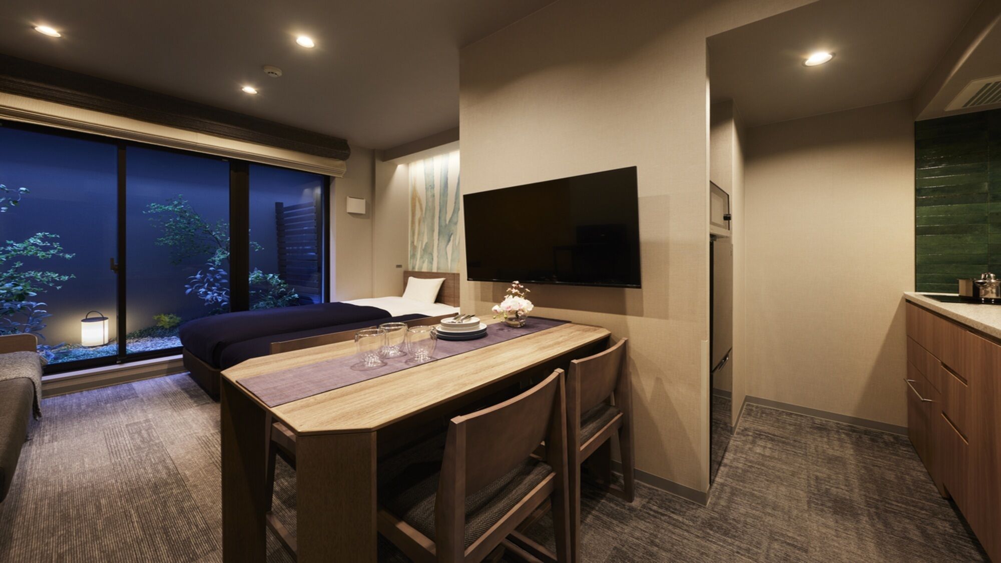 A modern hotel room with a large bed, a dining area with a table and chairs, a flat-screen TV mounted on the wall, and a view of a garden through the window.