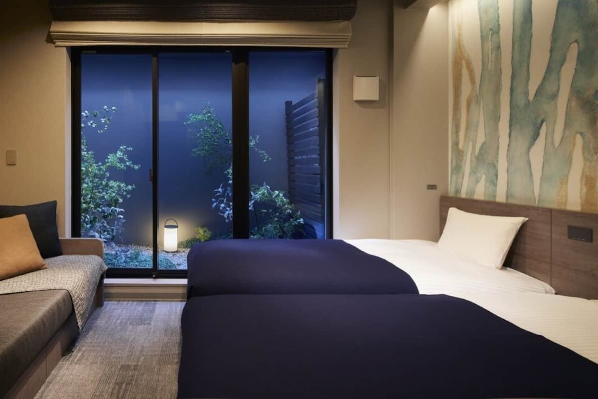 A hotel room with a bed, a sofa, a large window with a view of trees, and a wall with abstract art.