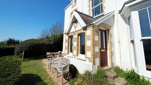 A two-story house with a white exterior and brown accents, a front door, and a small garden with a chair and table. - The Retreat - 5 Bedroom House - Horton (Swansea)