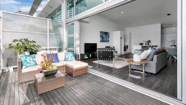 A modern living area with a large glass door leading to a balcony, wicker furniture, and a kitchen visible through the glass. - Princess Wharf w/ Balcony

 (Auckland)