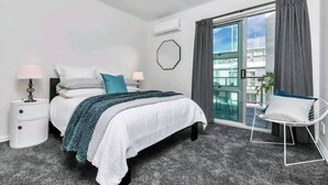 A modern bedroom with a bed, nightstands, a chair, and a view of a building through a glass door. - Princess Wharf w/ Balcony

 (Auckland)