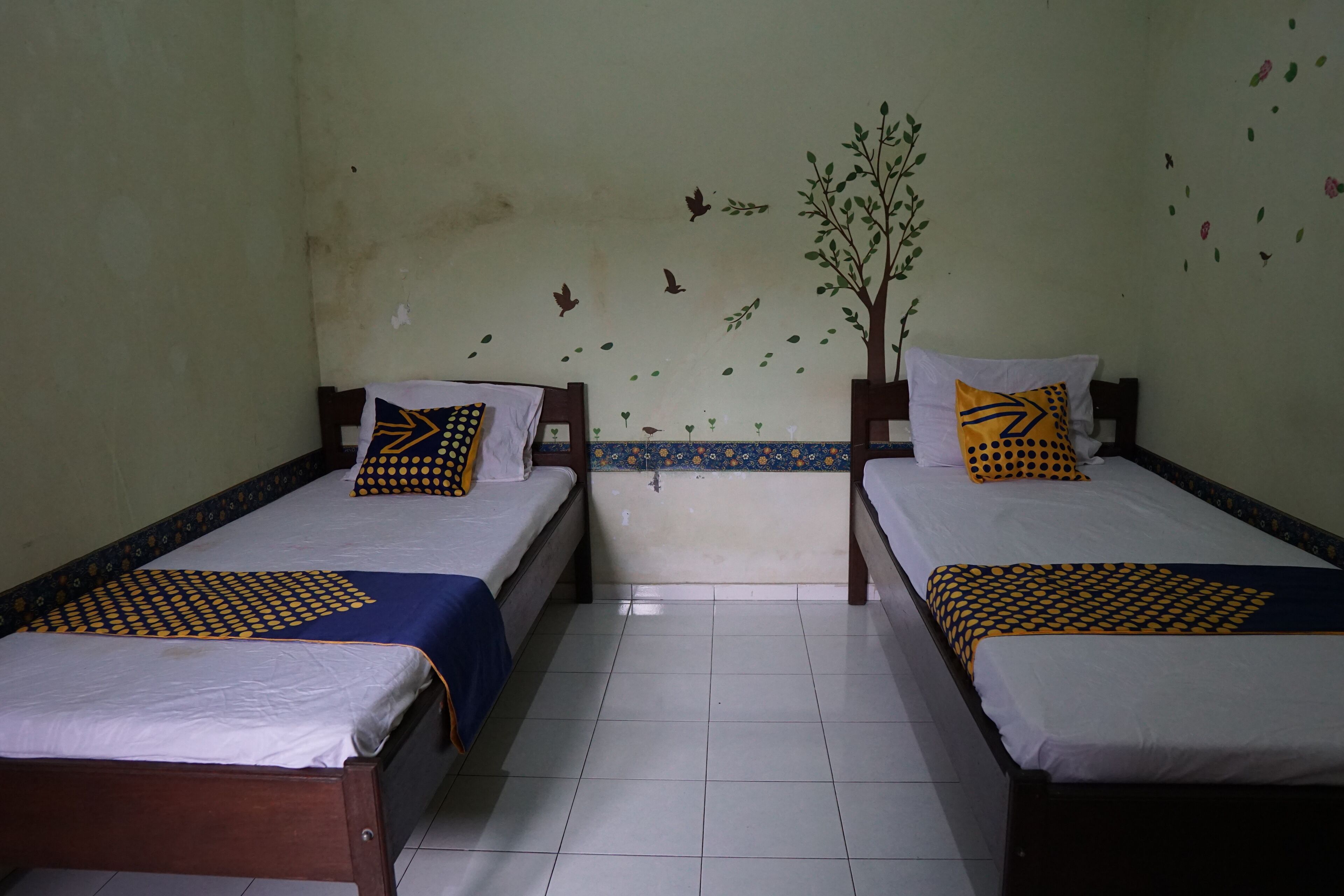 Une chambre avec deux lits simples, chacun recouvert d’une couvre-lit à motifs et agrémenté d’oreillers assortis. Les murs sont décorés de motifs floraux et d’arbres, et le sol est carrelé.