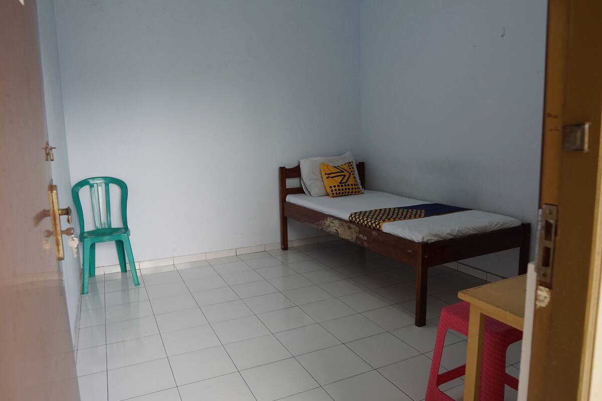 A small, single-bed room with a wooden frame, a green plastic chair, and a small table.
