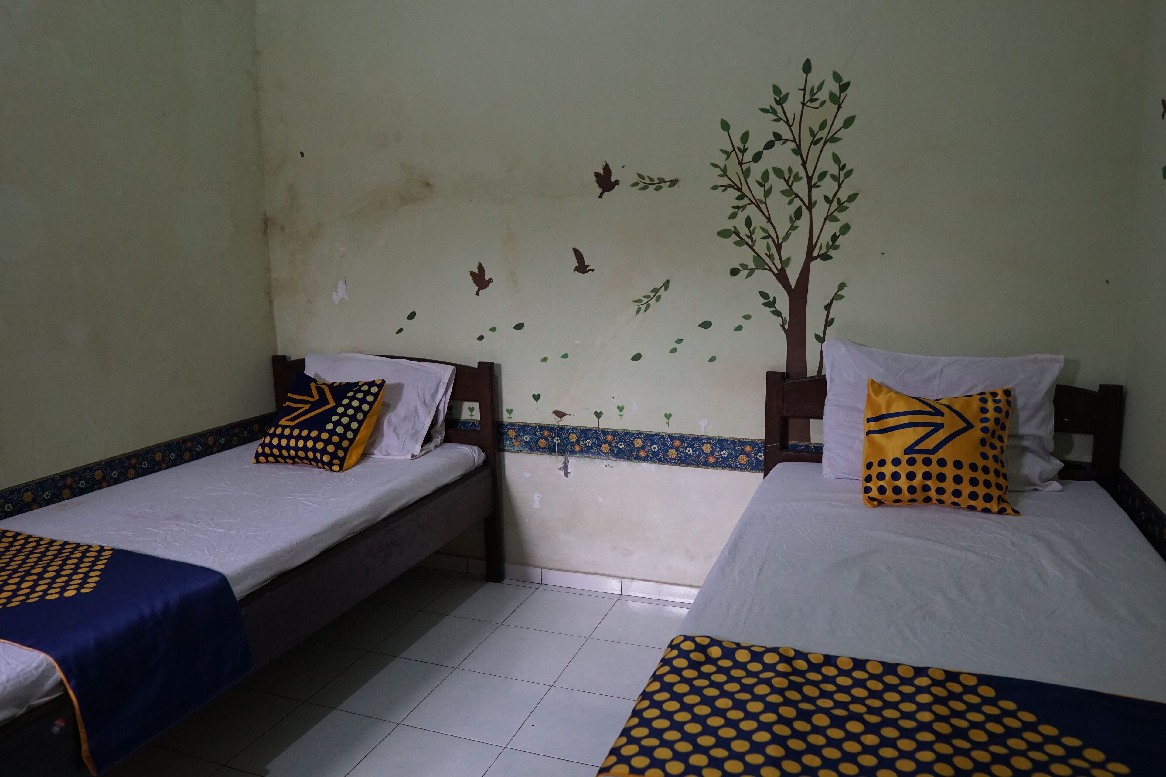 Two single beds with matching headboards and footboards, each with a white mattress and a blue and yellow patterned bedspread. The room has a tiled floor and a wall with a tree and bird decal.