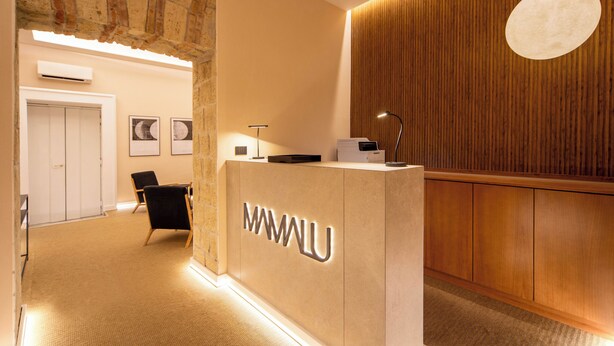 A reception area with a wooden reception desk, a black chair, and a wall-mounted light fixture. - Mamalu Studios and Suites (Naples)