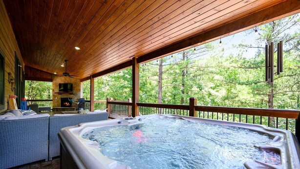 Een hoek met een jacuzzi, rieten meubels en een stenen openhaard, omgeven door een houten terras en met uitzicht op een bos.