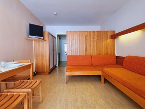 Quarto compacto com mesa de jantar de madeira, um sofá e uma televisão sobre um suporte.