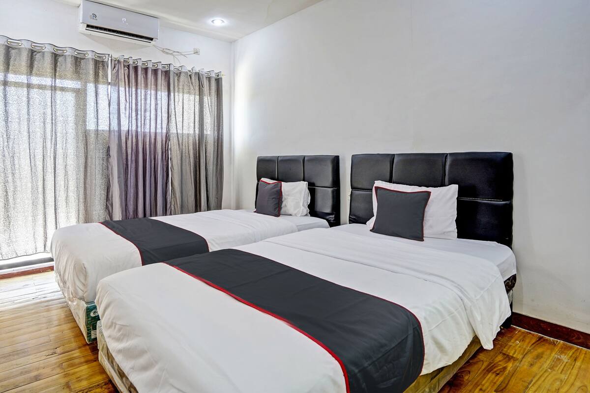 A hotel room with two beds, a wooden floor, a window with curtains, and an air conditioning unit on the wall.