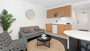 A compact living space with a sofa, coffee table, and a dining table. A kitchen area with wooden cabinets and a microwave is visible. - Perth's Premier King Deluxe (Perth)