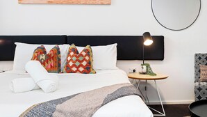 Ein ordentlich bezogenes Bett mit gemusterten Kissen, eine Nachttischlampe, ein kleiner Tisch mit Pflanze und ein an der Wand befestigter Spiegel.