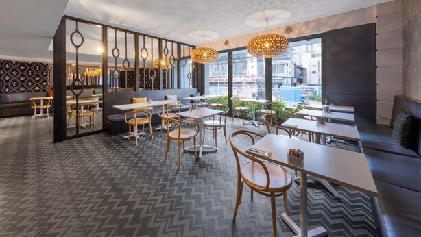 En modern restaurang med ett golv i sicksackmönster, trästolar och träbord samt en svart avskärmning med cirkelformade utskärningar.