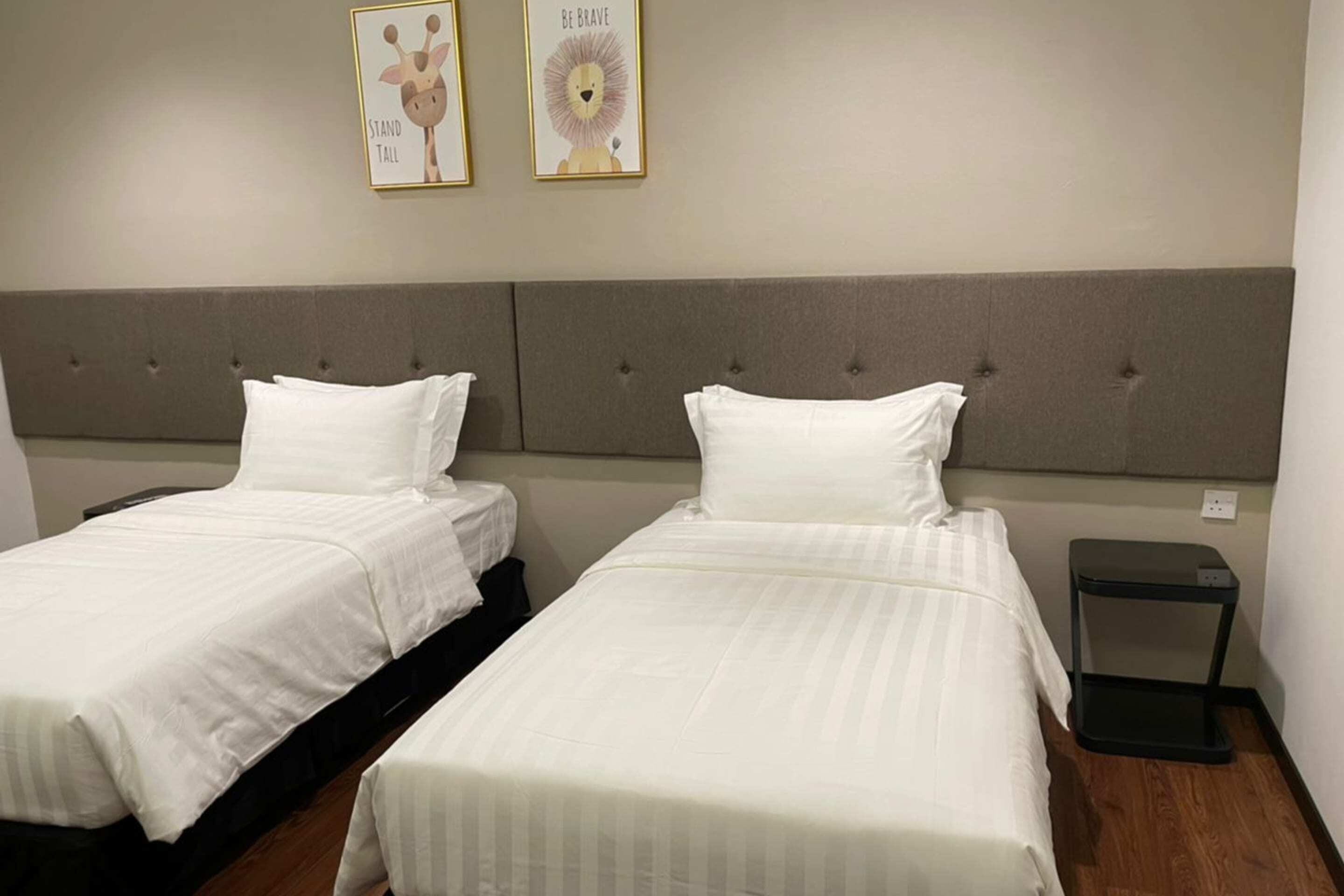 A hotel room with two beds, each with white bedding and a grey headboard. There is a small black nightstand beside one bed.