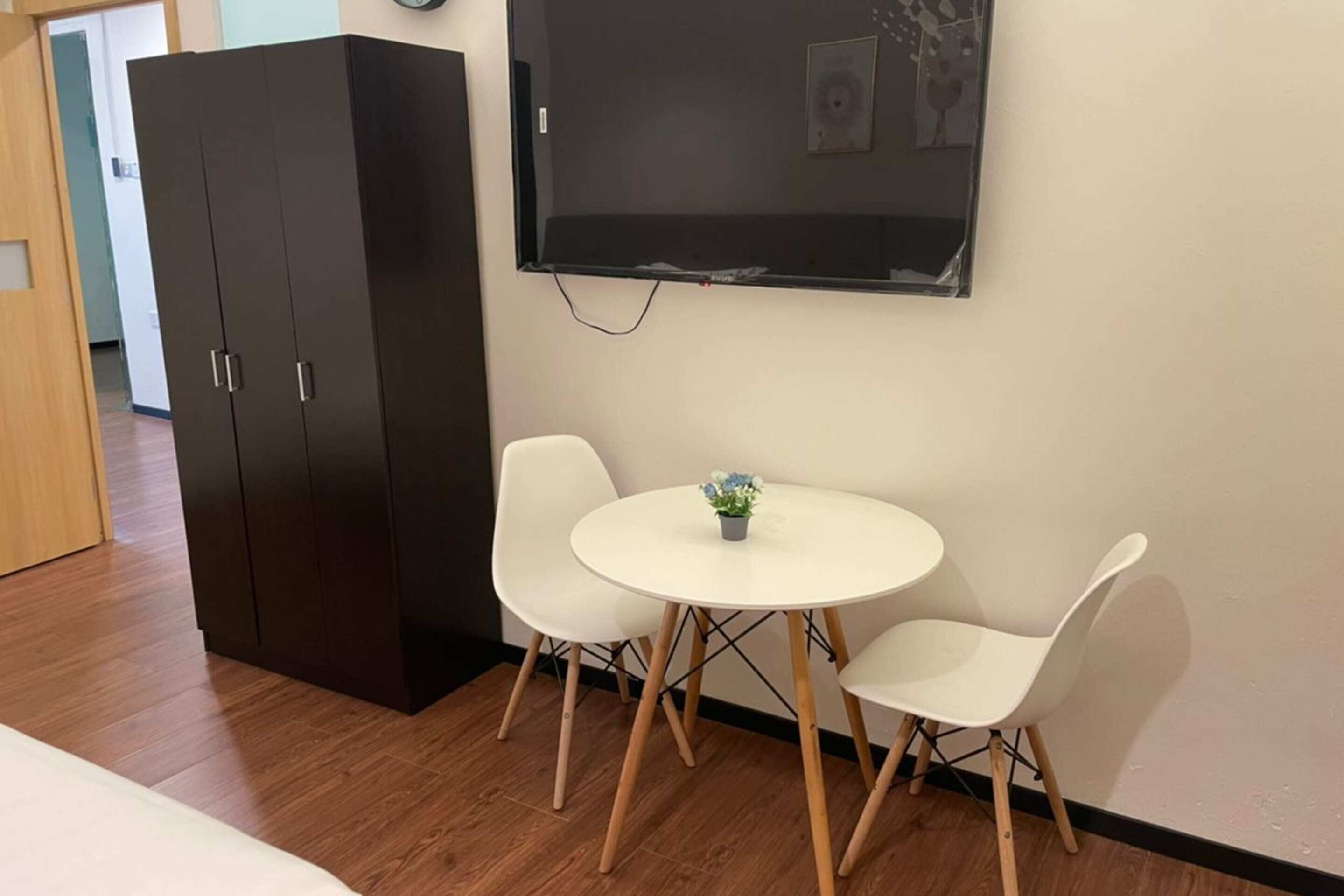 A room with a large dark wardrobe, a round table with two white chairs, and a wall-mounted TV.