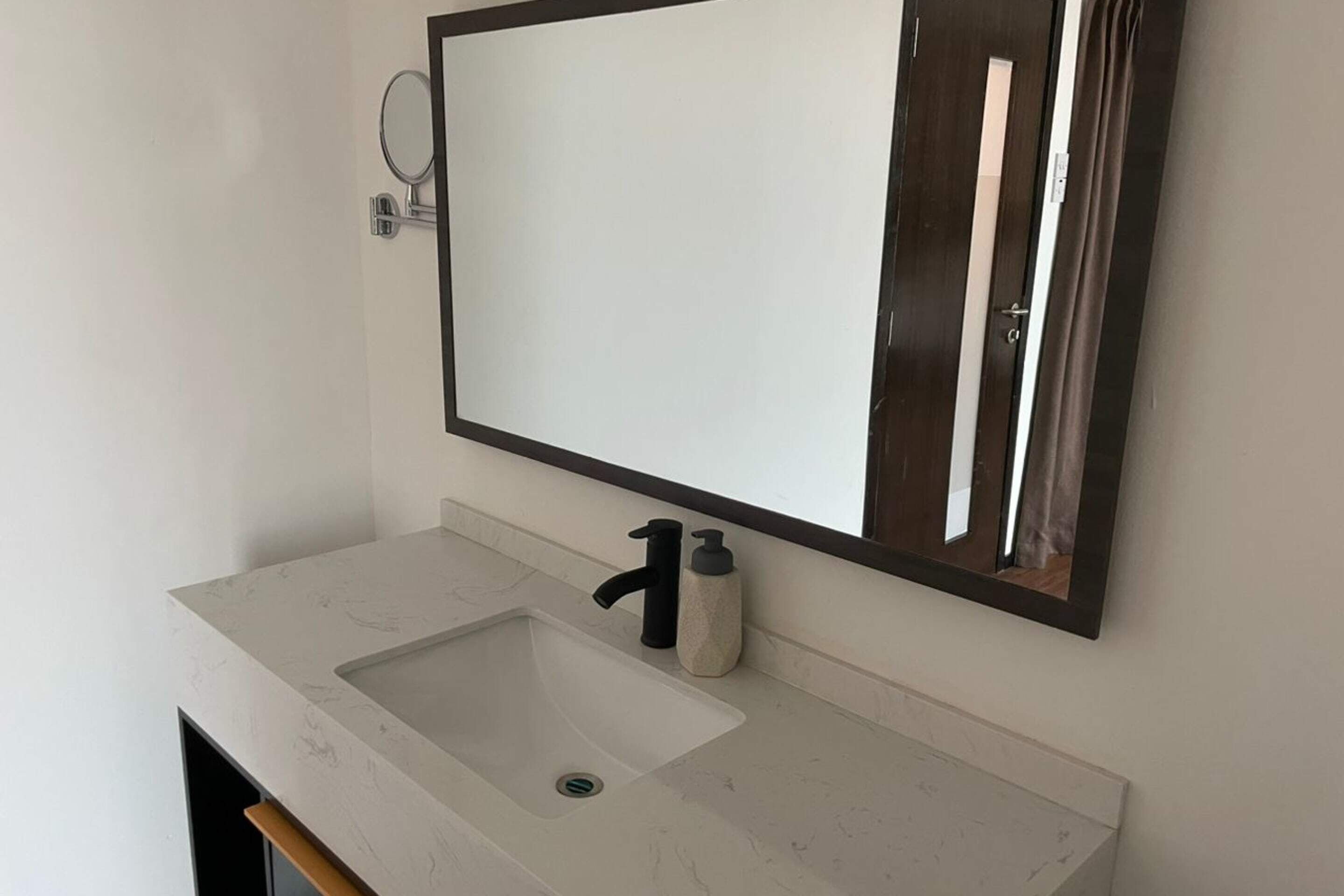 Une salle de bain avec un lavabo blanc, un robinet noir, un distributeur de savon et un miroir avec un cadre en bois.