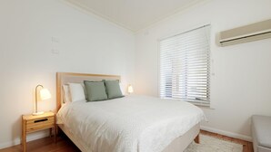 Une chambre à coucher comprenant un lit, deux lampes de chevet, une fenêtre avec stores et un appareil de climatisation.