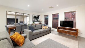 Ein modernes Wohnzimmer mit grauem Sofa, einem Fernseher auf einem Holzschrank und einem Essbereich mit Tisch und Stühlen.