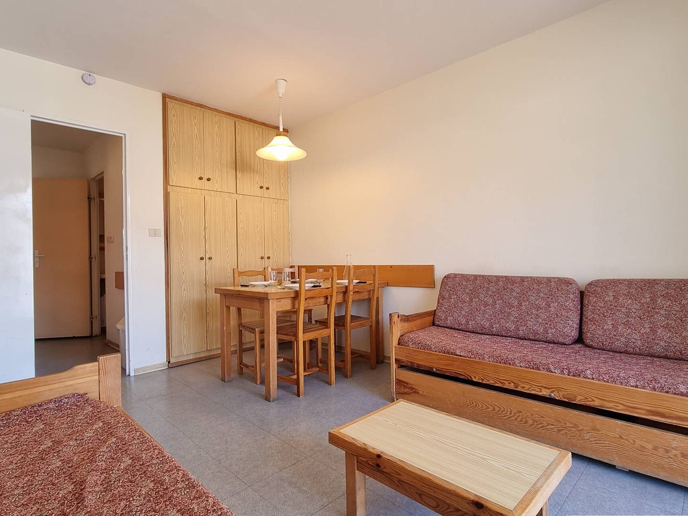 Een kleine, goed verlichte kamer met een houten eettafel en stoelen, een bank en een bijzettafeltje.