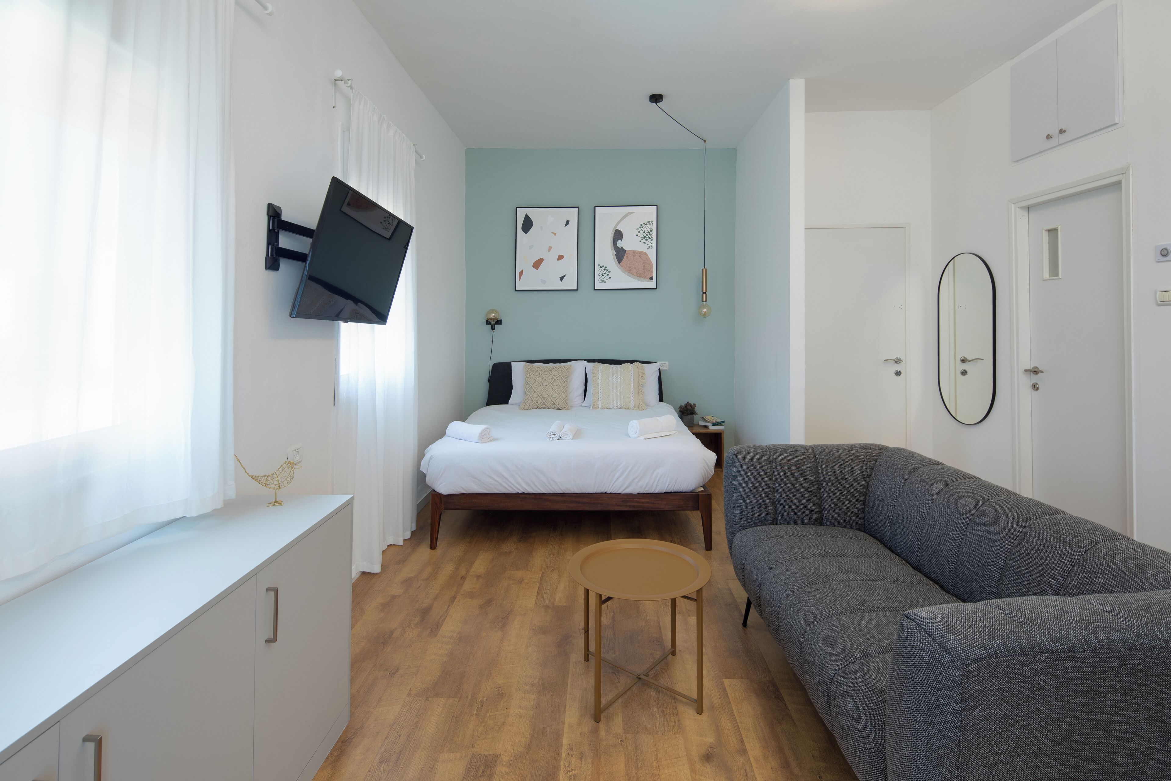 A bedroom with a bed, a grey sofa, a small round table, and a TV mounted on the wall.