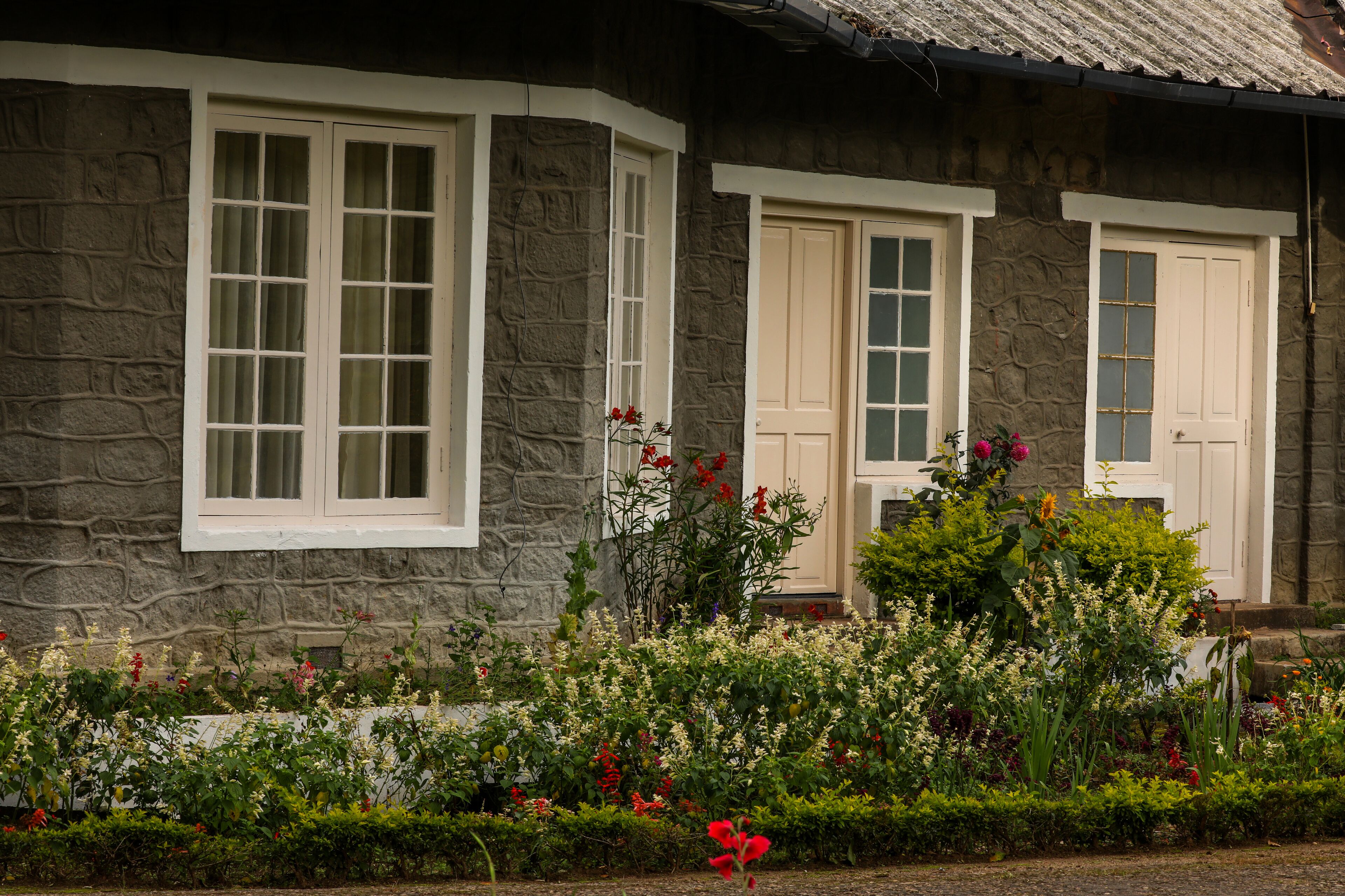 A stone building with two white-framed windows and doors, surrounded by a well-maintained garden with various flowers.