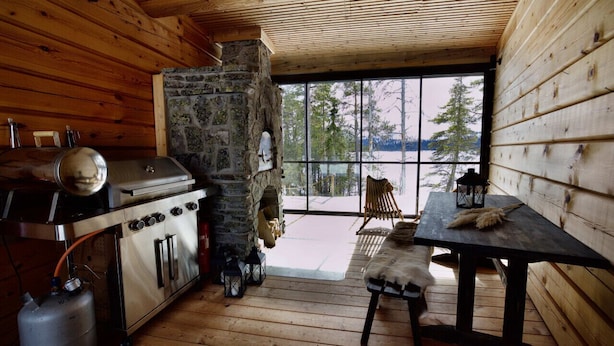 Ein Interieur einer Holzhütte mit Kamin aus Stein, Holzwänden, einem Esstisch und Blick auf Bäume durch große Fenster.