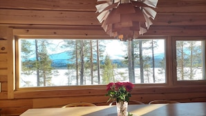 Ein Holzblockhaus-Interieur mit einem Esstisch, einer Vase mit Blumen und Blick auf eine verschneite Landschaft durch große Fenster.