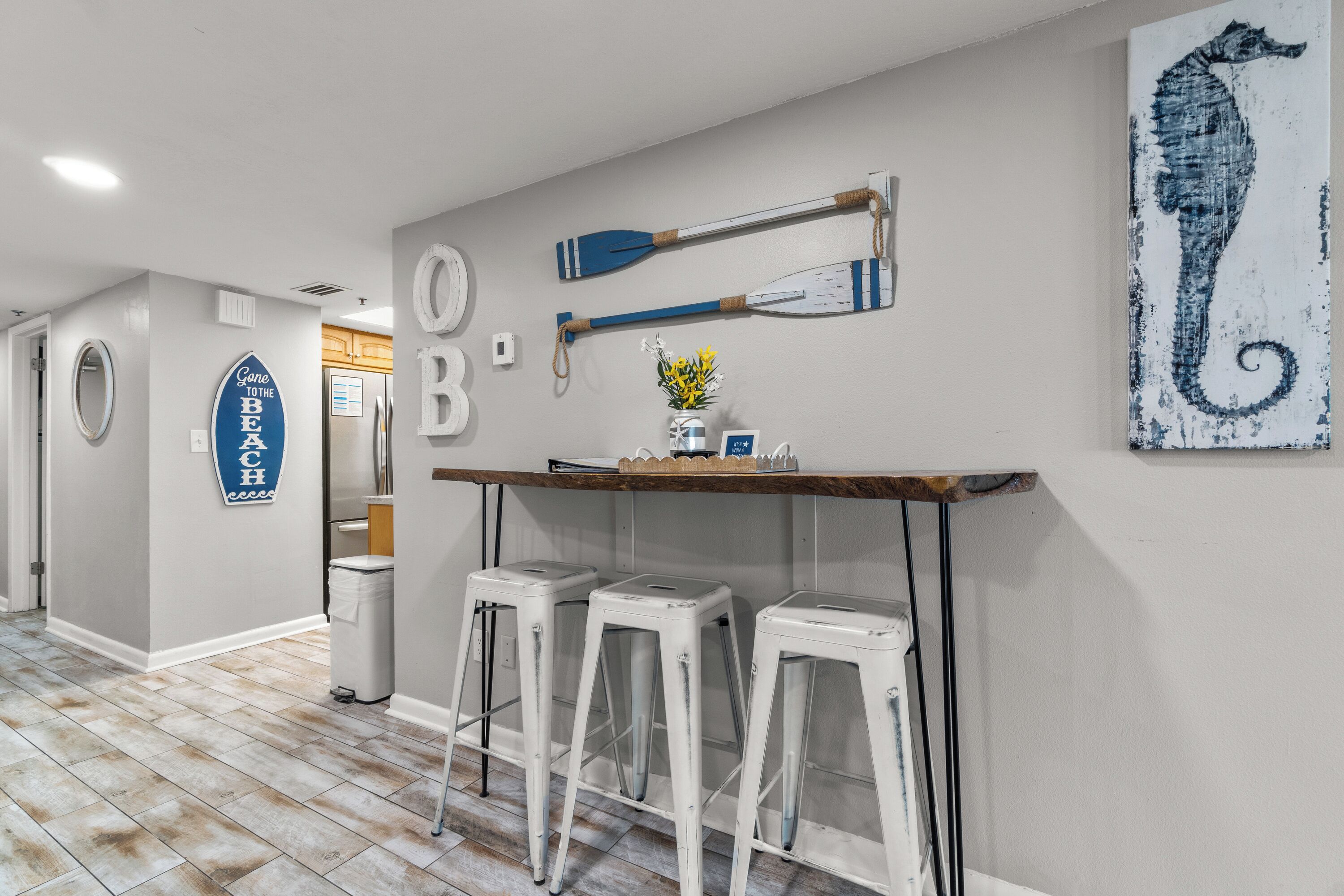 A hallway with a wooden floor, white walls, and a wooden bar counter with stools. There are two potted plants on the counter.