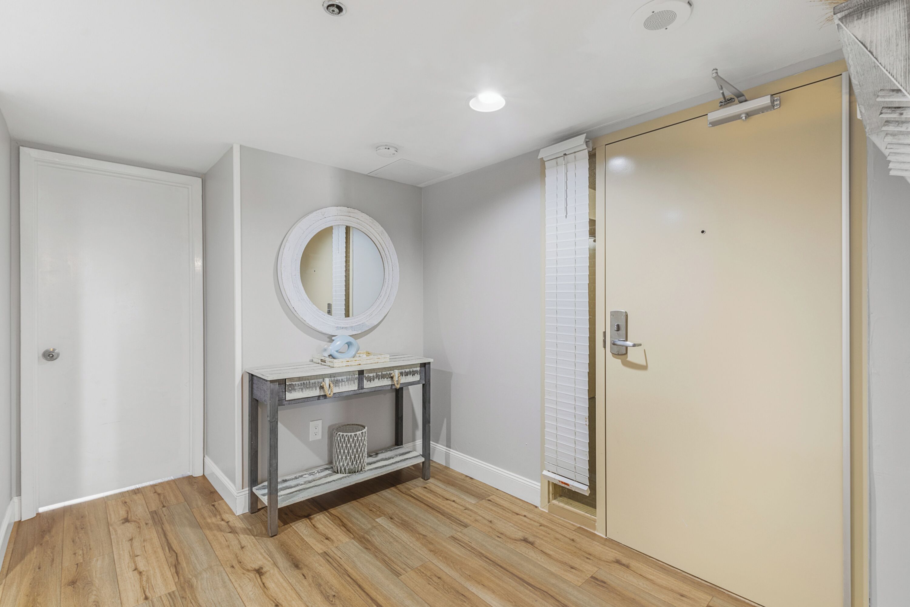 Une pièce avec un plancher en bois, une porte blanche, une console avec un miroir et une porte coulissante avec des stores.