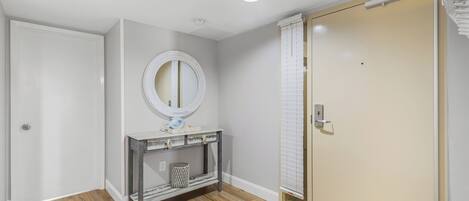 Une pièce avec un plancher en bois, une porte blanche, une console avec un miroir et une porte coulissante avec des stores.