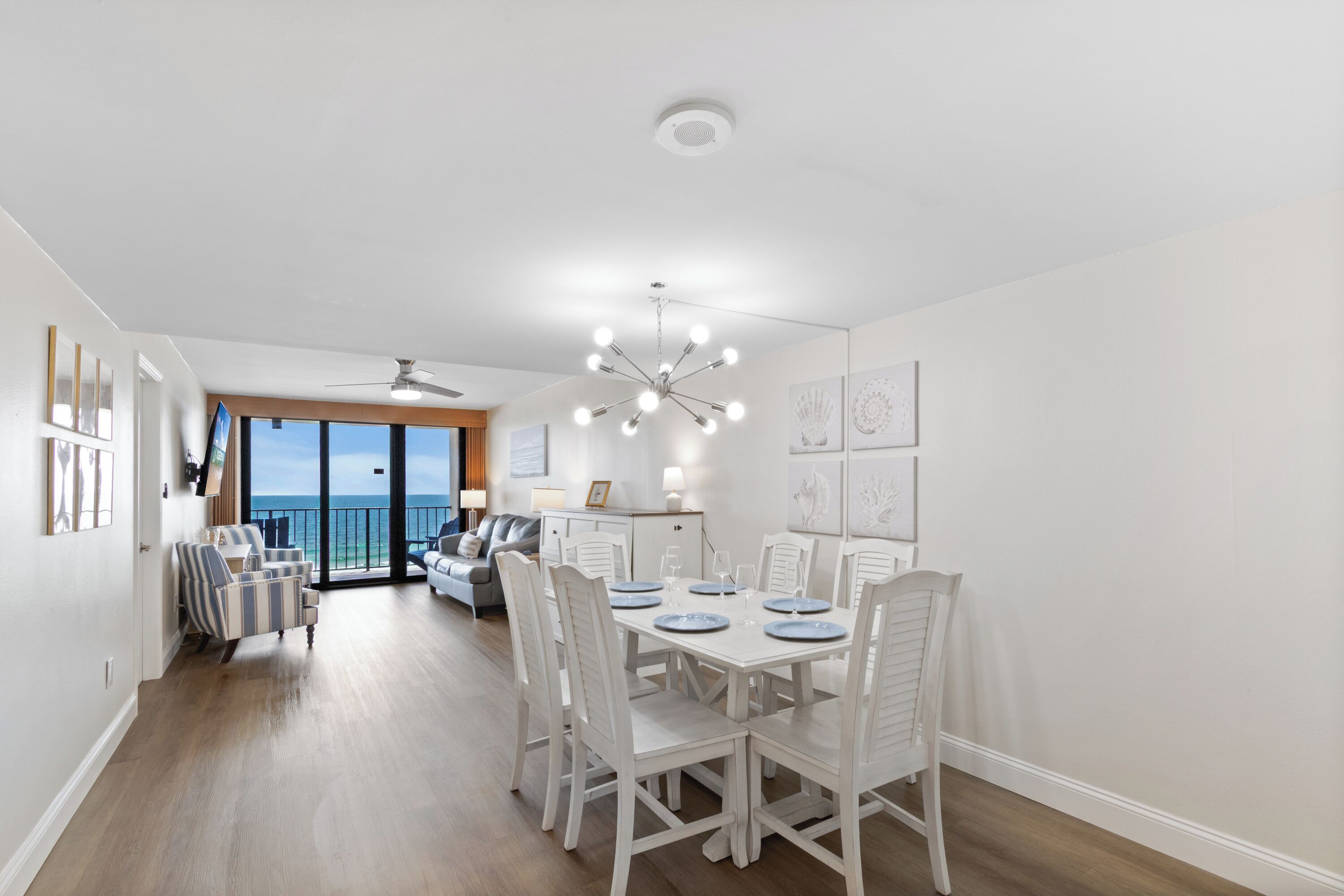 Un coin repas avec une table blanche dressée pour six personnes, des chaises blanches, un ventilateur de plafond et une vue sur l’océan grâce à de grandes fenêtres.