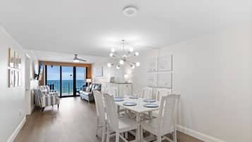 A dining area with a white table set for six, white chairs, a ceiling fan, and a view of the ocean through large windows.
