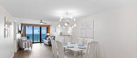 Un coin repas avec une table blanche dressée pour six personnes, des chaises blanches, un ventilateur de plafond et une vue sur l’océan grâce à de grandes fenêtres.