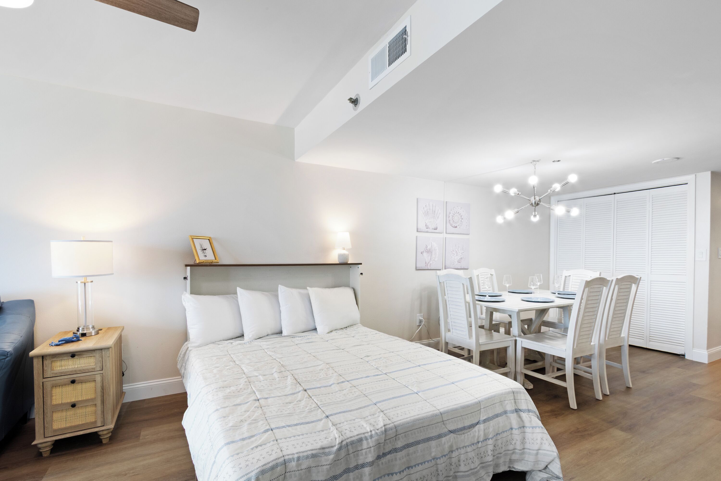 A bedroom with a bed, bedside table, lamp, and a dining area with chairs and a table.