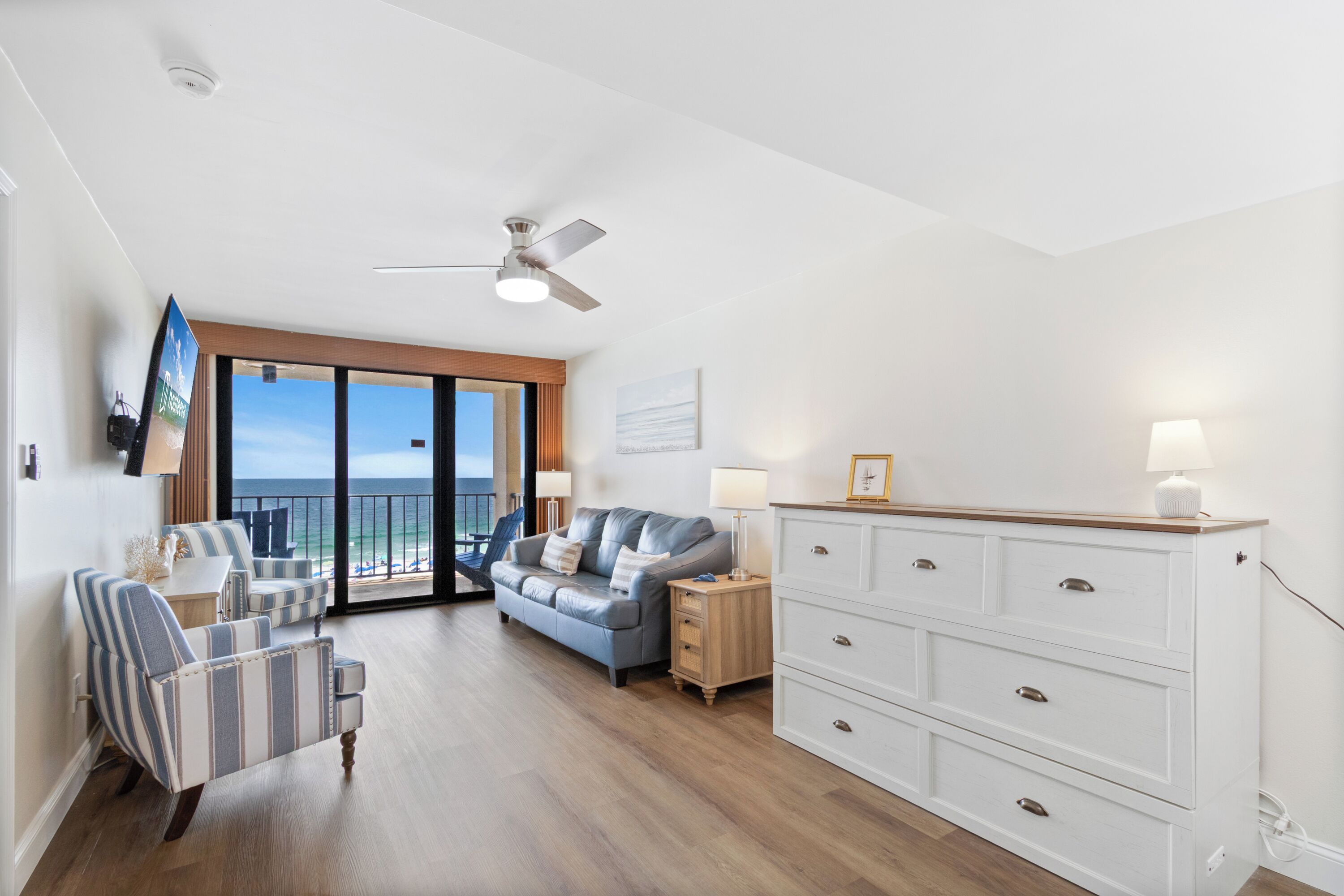 A living room with a sofa, armchairs, a TV, a ceiling fan, a wooden cabinet, and a view of the ocean.